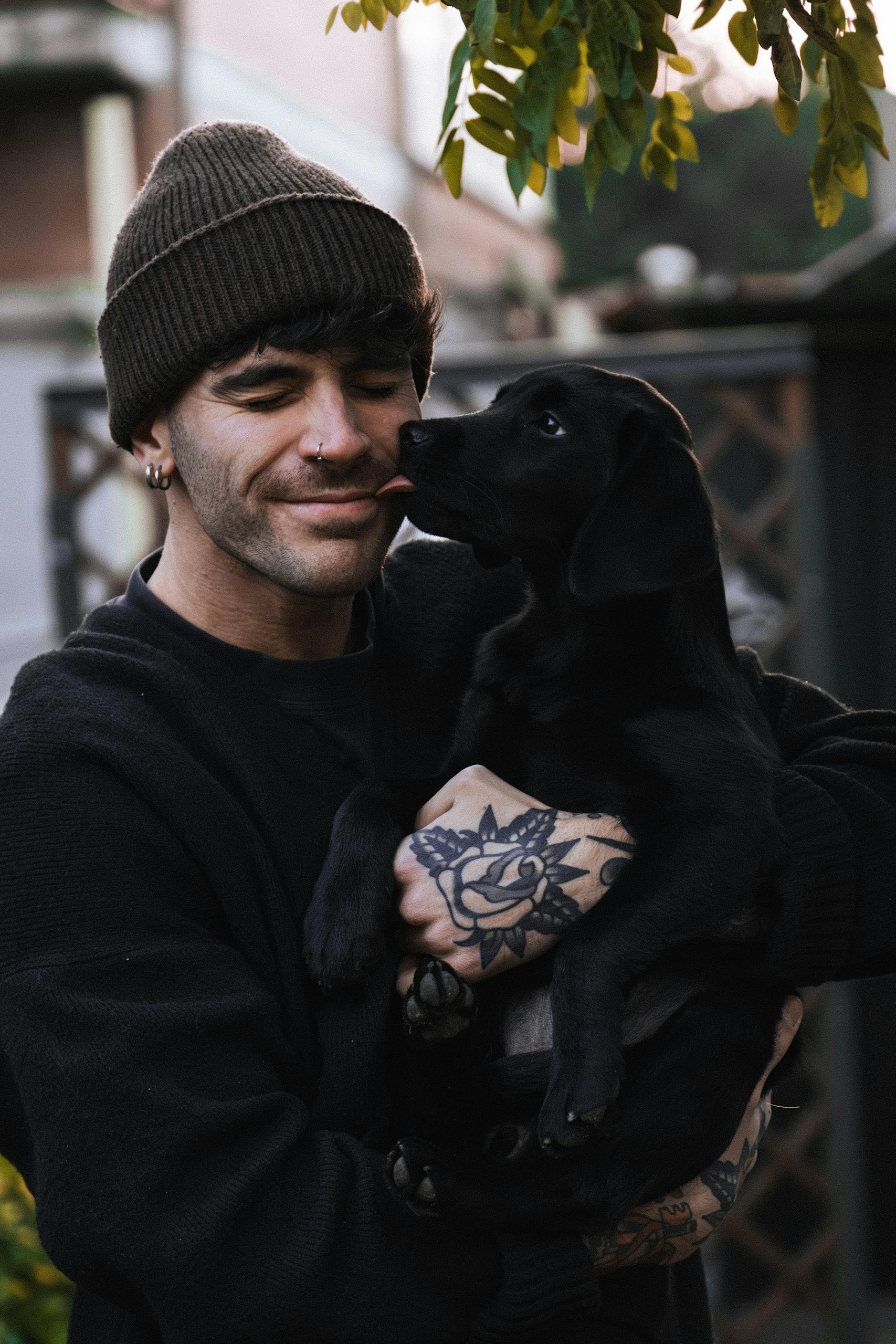 Smiling man with tattoos holding a playful black Labrador puppy outdoors.